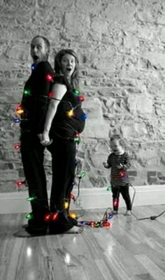 a man and woman standing in front of a wall with christmas lights on it while a small child stands next to them