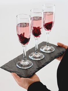 three glasses of wine are sitting on a slate serving tray with red rose petals in them