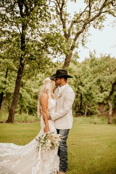 a bride and groom are standing in the grass together, dressed in cowboy attire with white flowers