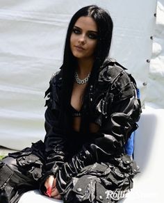 a woman sitting on top of a white bench wearing black clothes and jewelry, with her hands in her pockets