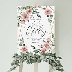 a welcome sign with pink flowers and greenery on an easel for a wedding