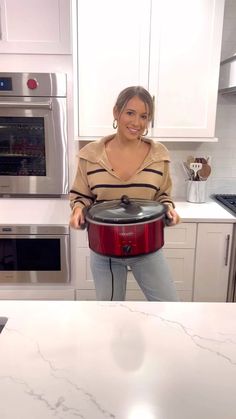 a woman standing in a kitchen holding a red crock pot with the lid open