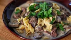 a red bowl filled with pasta and meat covered in mushroom sauce on top of a wooden table