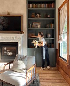 a woman standing in front of a fireplace holding a vase filled with flowers and looking out the window