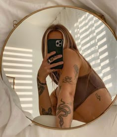 a woman with tattoos taking a selfie in front of a round mirror while holding her cell phone