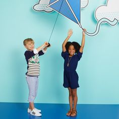 two children are holding up a kite in front of a blue wall with clouds on it