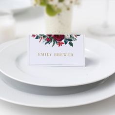 a place card is placed on a white plate next to a vase with red flowers