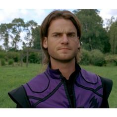 a man with long hair wearing a purple shirt and black tie standing in front of a green field