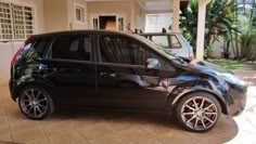 a small black car parked in front of a house next to a driveway with palm trees