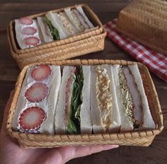 a hand holding a basket filled with sandwiches on top of a wooden table next to another basket