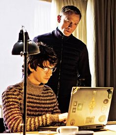 two people standing in front of a laptop computer on a desk next to a lamp