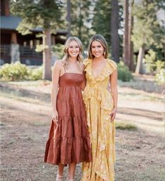 two women standing next to each other in front of some trees and grass with one wearing a brown dress