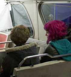 two people sitting on a bus with pink hair and green sweaters looking out the window