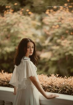 a woman in a white dress standing on a ledge looking at the camera with trees and bushes behind her