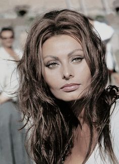 a woman with long brown hair standing in front of a group of people and looking at the camera