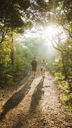 two people walking down a dirt path in the woods on a sunny day with sun shining through the trees