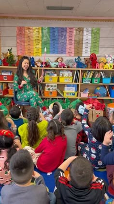 a woman standing in front of a classroom full of children sitting on the floor and talking to them