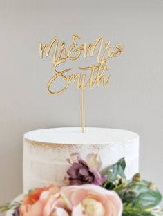 a white cake topped with a gold mr and mrs cake topper next to flowers