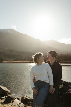 a man and woman standing next to each other on rocks near water with mountains in the background