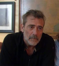 an older man with grey hair and beard sitting in front of a framed photo on the wall