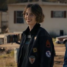 a woman standing in front of a house wearing a black jacket with patches on it