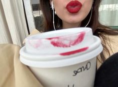 a woman with red lipstick holding a white cup in front of her face while wearing ear buds