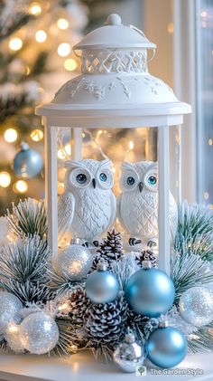 two owls sitting on top of a white lantern surrounded by christmas ornaments and pine cones