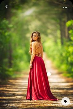 a woman in a red dress is standing on a dirt road with trees behind her