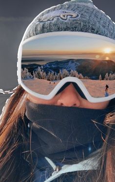 a woman wearing ski goggles and a hat with the sun setting in the background