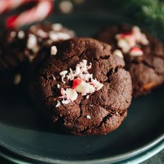 three chocolate cookies with white and red candy canes on a black plate next to peppermint sprigs