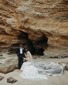 a bride and groom standing in front of a rock formation on the beach with their veil blowing in the wind