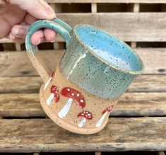 a hand holding a blue and brown coffee mug on top of a wooden bench with mushrooms painted on it