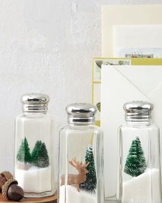 two glass jars filled with snow and trees on top of a wooden table next to an envelope