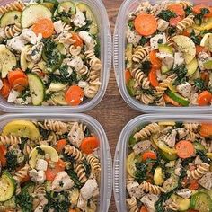 four plastic containers filled with pasta salads on top of a wooden table next to carrots and cucumbers