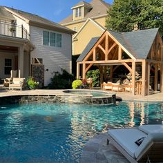 an outdoor pool surrounded by patio furniture and water features in front of a house with large windows