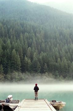 a man standing on a dock next to two boats in the water near a forest