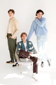 three people standing and sitting on a chair in front of a white wall with one person wearing a blue sweater