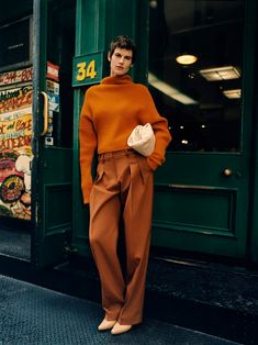 a woman standing in front of a green door wearing an orange sweater and brown pants