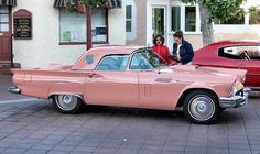 two people standing next to a pink car in front of a white and red building