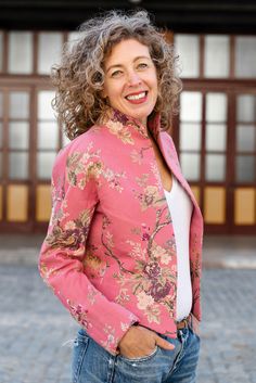 a woman with curly hair wearing a pink jacket and jeans standing in front of a building