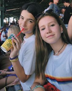 two young women sitting next to each other at a baseball game, one drinking a drink and the other holding a straw in her hand