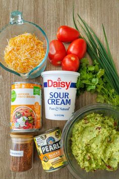 the ingredients to make guacamole are displayed on a wooden surface with green onions, tomatoes, cheese and sour cream