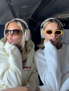 two women wearing headphones and sunglasses sitting in the back of an airplane with their hands on their chins