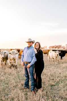 a man and woman standing in the middle of a field surrounded by cows at sunset