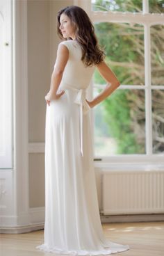 a woman standing in front of a window wearing a white dress with a bow at the waist