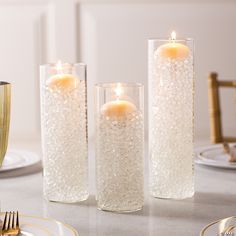 three glass vases filled with water and lit candles on a dining room table set for two