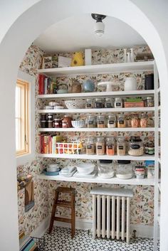 an arched doorway leads into a kitchen with wallpapered walls and white shelves filled with dishes