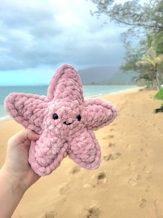 a hand holding up a pink crocheted starfish on a sandy beach next to the ocean