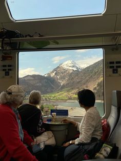 three people are sitting on a train looking out the window at mountains and lake in the distance