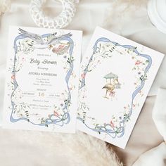 two wedding cards on top of a table with feathers and flowers around them, sitting next to each other